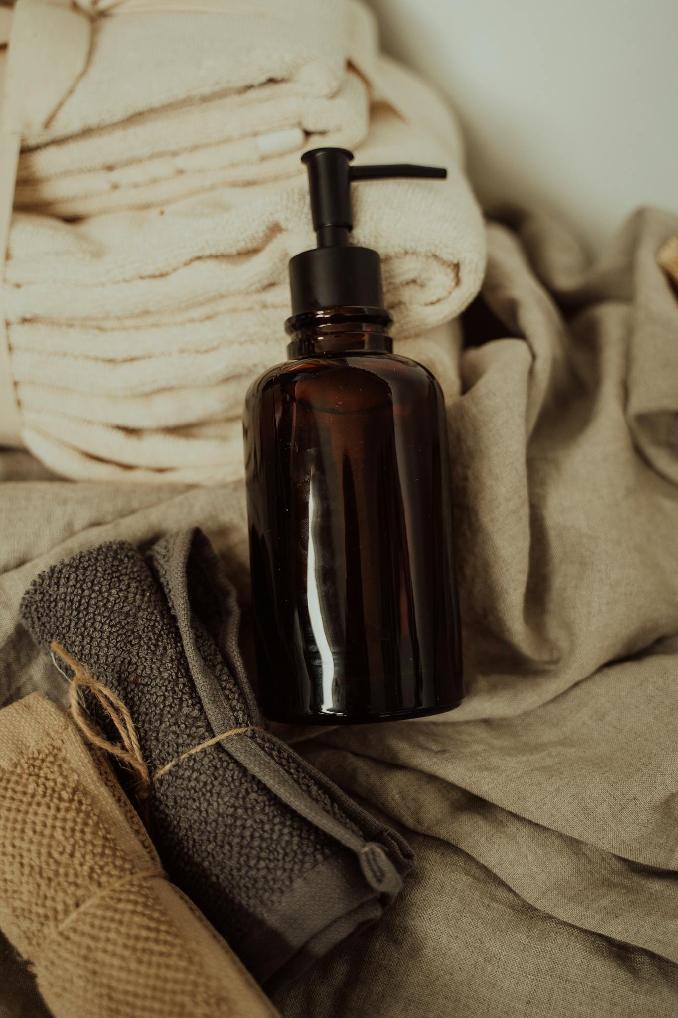 Close-up of a dark amber pump bottle with folded face towels in a neutral setting.